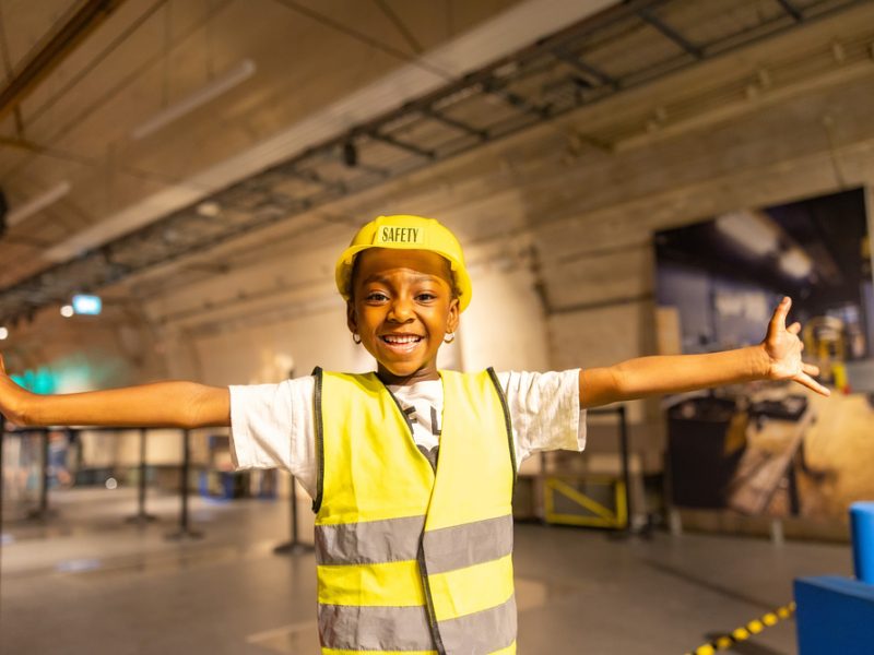 A girl wearing a high-vis vest and hard hat opens her arms and looks at the camara with big smile