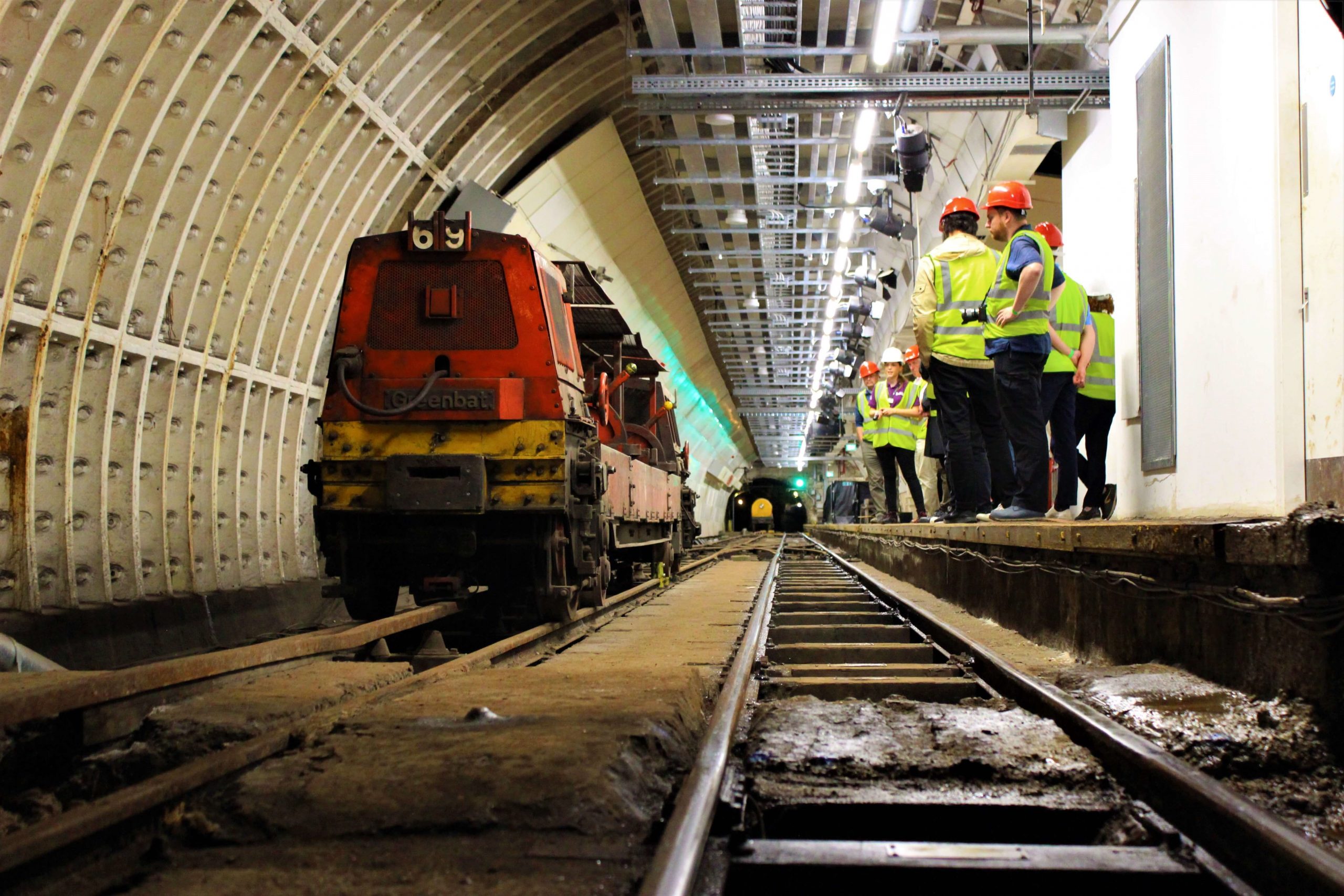Group members wearing helmets and high-visibility jackets stand on underground platform next to an old Mail Rail train.