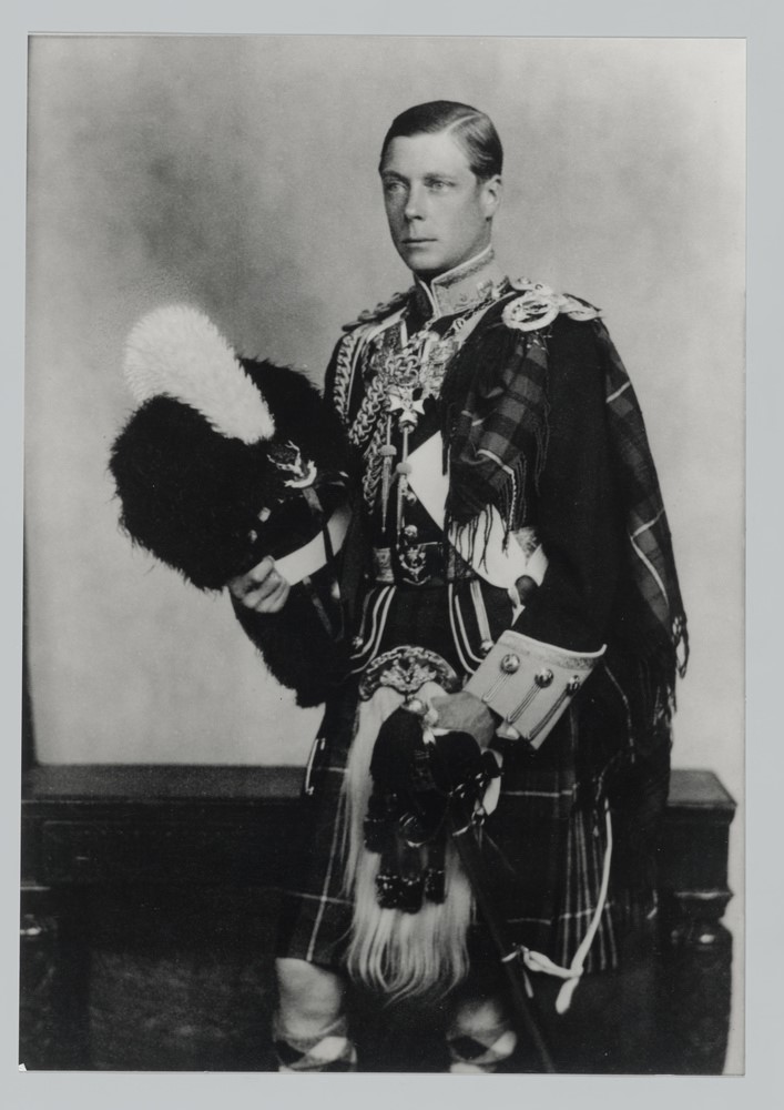 The King wears a very ornate outfit in this black and white photograph. He is wearing a kilt, lots of medals and holding furry hat.