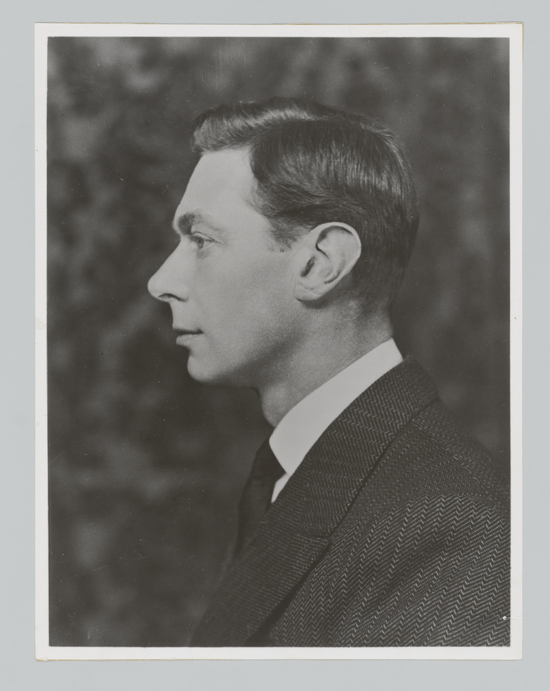 The king wears a pinstriped suit, white shirt and black tie in this black and white photograph. He is facing the left so you can only see his left-side profile.