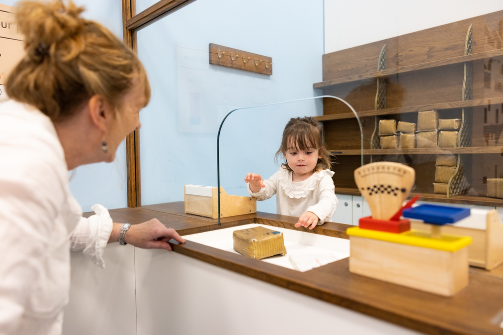 A small girl and woman play at a miniature postal counter, using toy scales and miniature parcels.