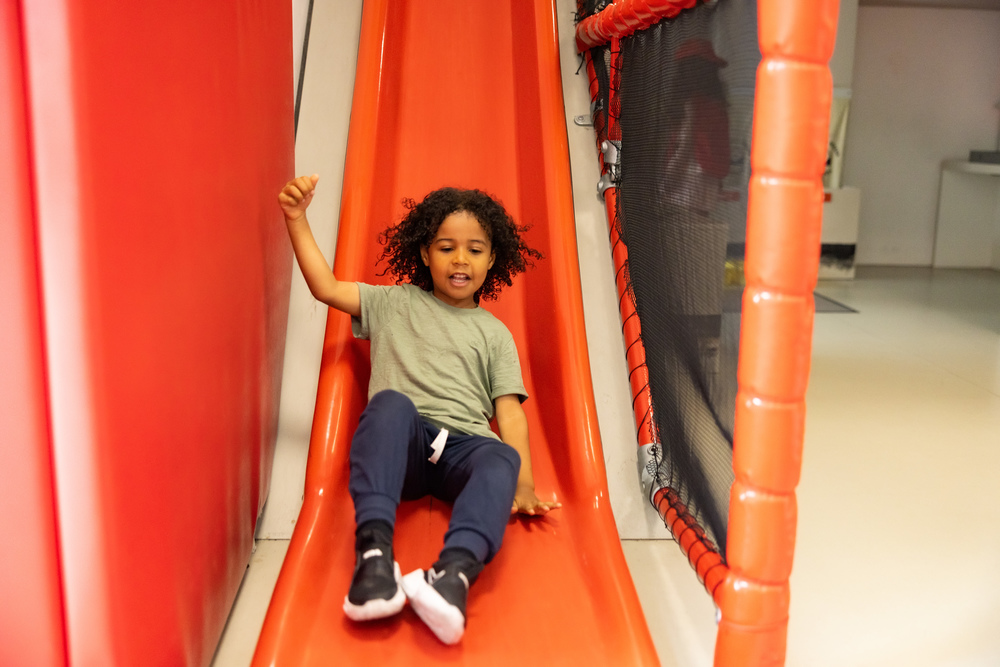 A boy with a light green t-shirt and blue trousers plays on a red slide.
