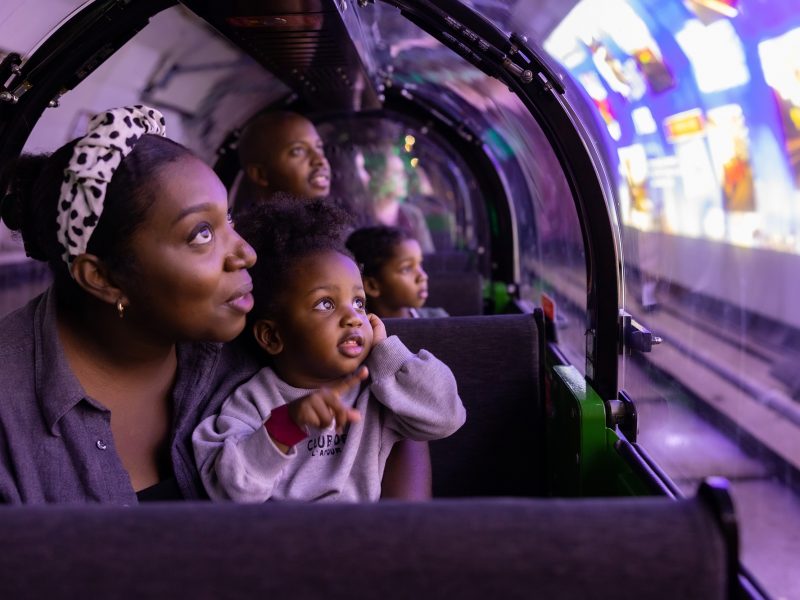 A mother and child sitting in the carriage of a miniature train. They are looking out the window at a video being projected onto the walls of the train platform.