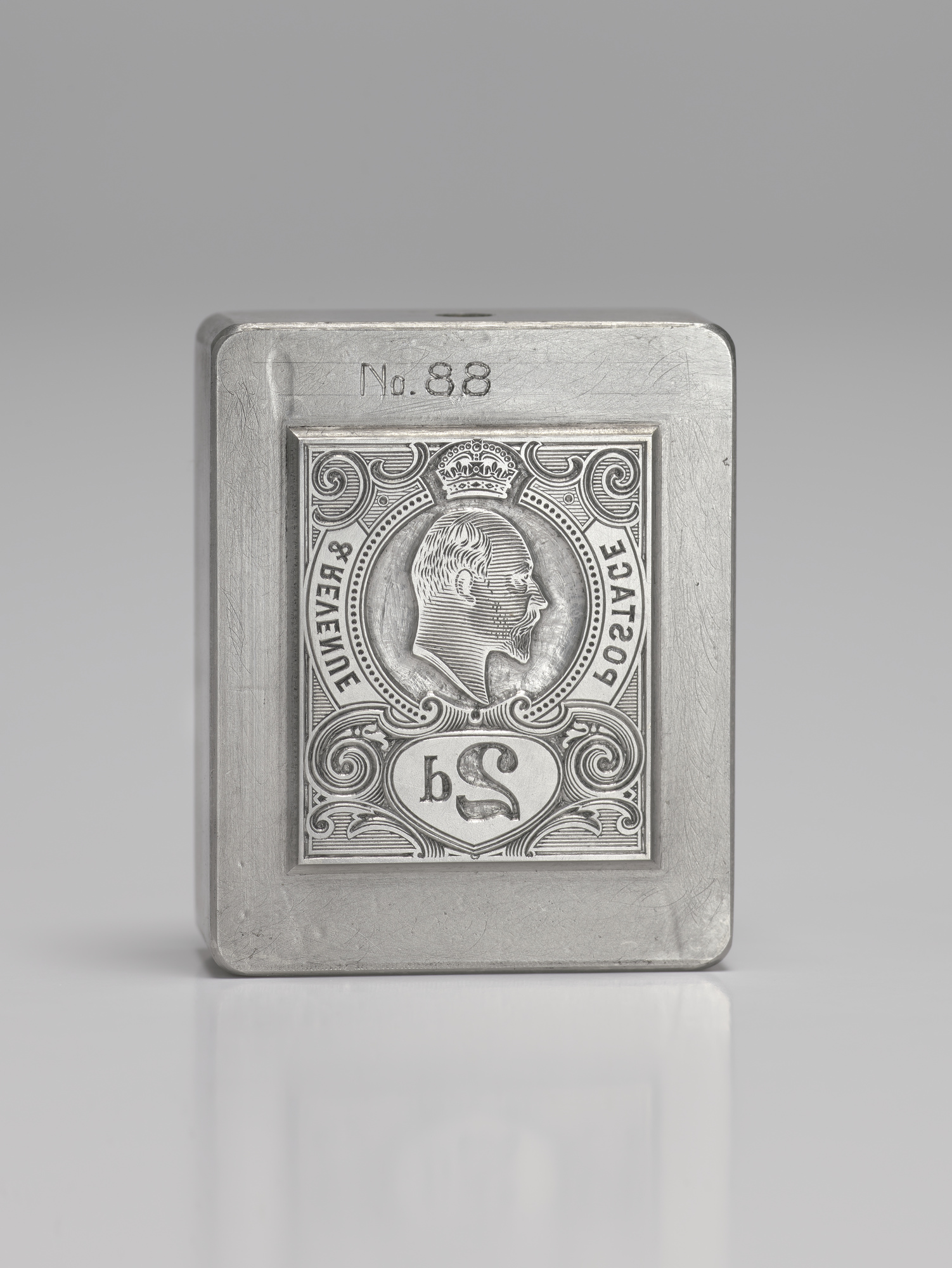 A metal die cube, showing a stamp design in reverse - an image of a king in a crown is in the middle, with the letters '2d' below this.