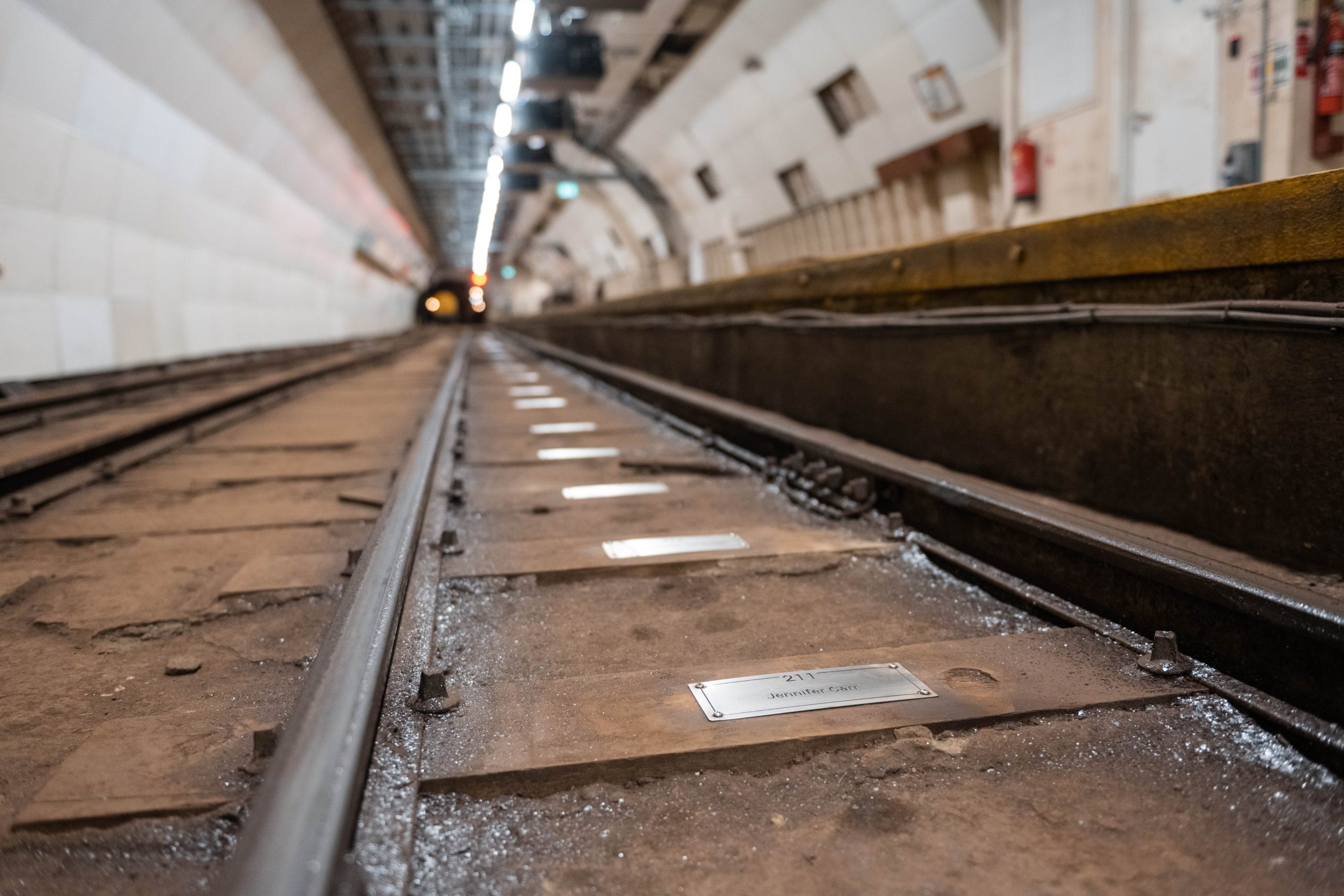 A closeup view of several silver coloured metal plaques, fixed to wooden sleeper rails on the floor of the Mail Rail tunnel.