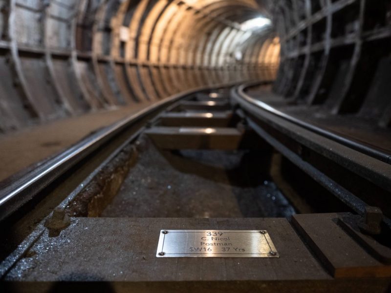 A closeup view of a silver coloured metal plaque, fixed to a wooden sleeper rail on the floor of the Mail Rail tunnel.