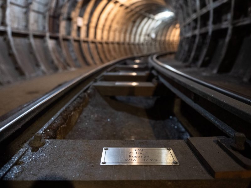 A closeup view of a silver coloured metal plaque, fixed to a wooden sleeper rail on the floor of the Mail Rail tunnel.