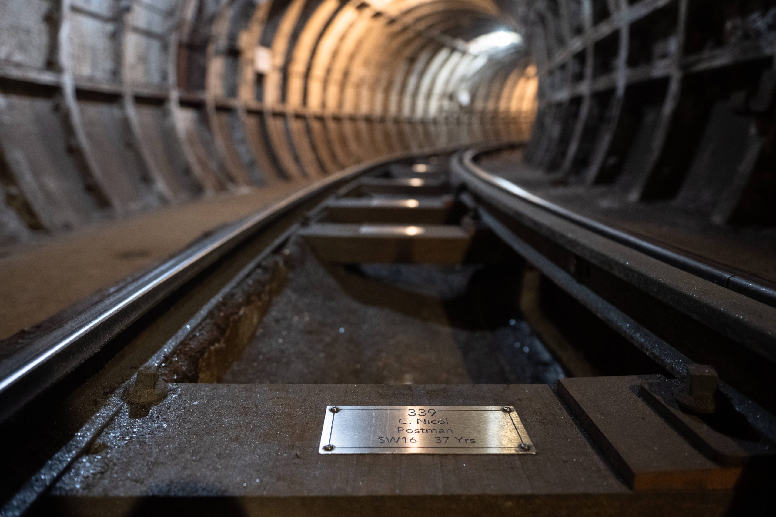 A closeup view of a silver coloured metal plaque, fixed to a wooden sleeper rail on the floor of the Mail Rail tunnel.