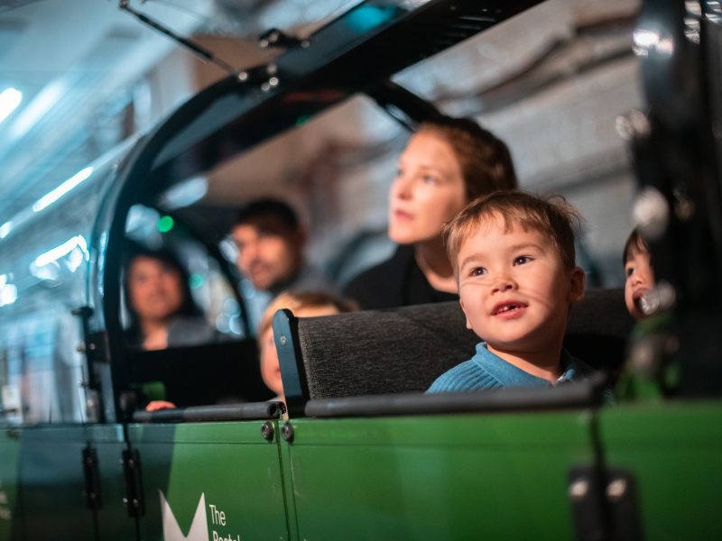 A smiling young boy rides Mail Rail with his mum sat behind him. You can see the green Mail Rail carriage is full with families.