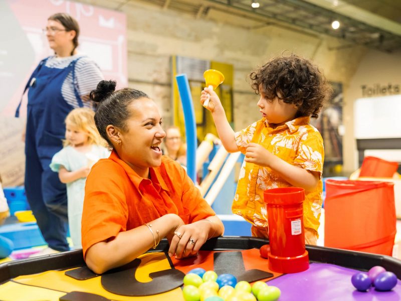 A mum laughs with her son as he rings a orange bell. They both wear bright coloured clothing.