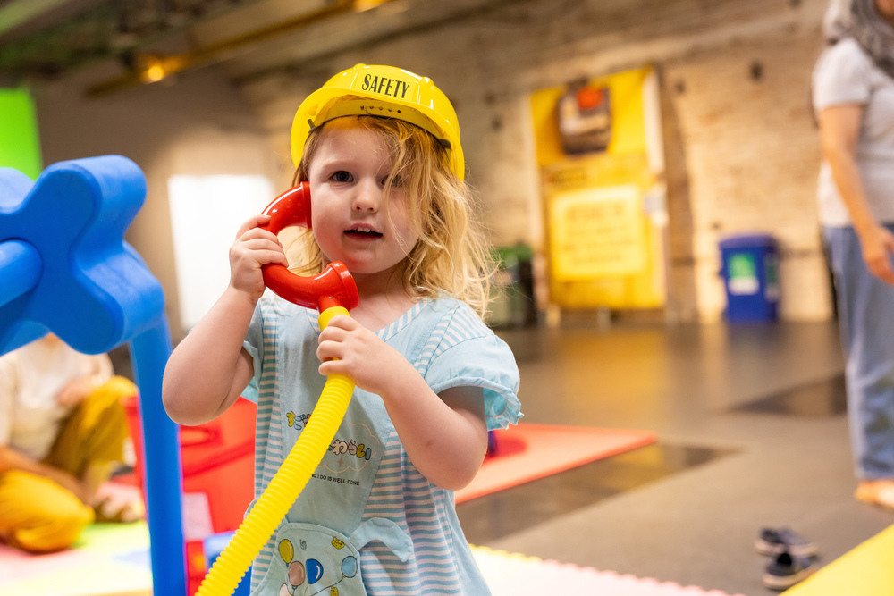 A young girl in a gingham dress makes a pretend phone call in our colourful Construction Zone. She wears a yellow hard hat.