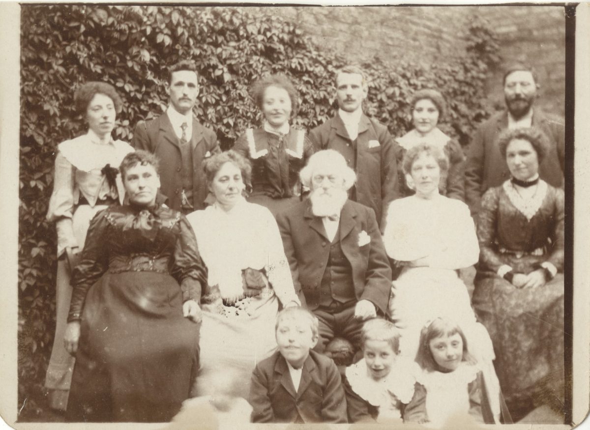 Everyone in this photograph looks very Victorian. Only the 3 small children sat at the bottom of the picture are smiling. The two rows of adults, a mixture of men and women look stoic. Not all look directly at the camera. A few of the people are quite blurry. Everyone has dark hair apart from the elderly gentle man sat in the middle of the second row, two women either side of him. He has white hair and a white bushy beard. The top row of people stand boy, girl, boy, girl, boy girl. 