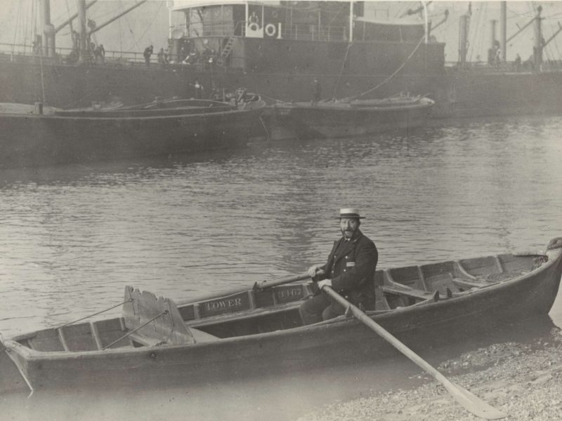 This is a very old, black and white photograph of a middle-aged bearded man sat in a long wooden boat, known as a skiff. He wears a dark uniform and a straw boaters hat. He has a long wooden oar in each hand and is looking directly at the camera.