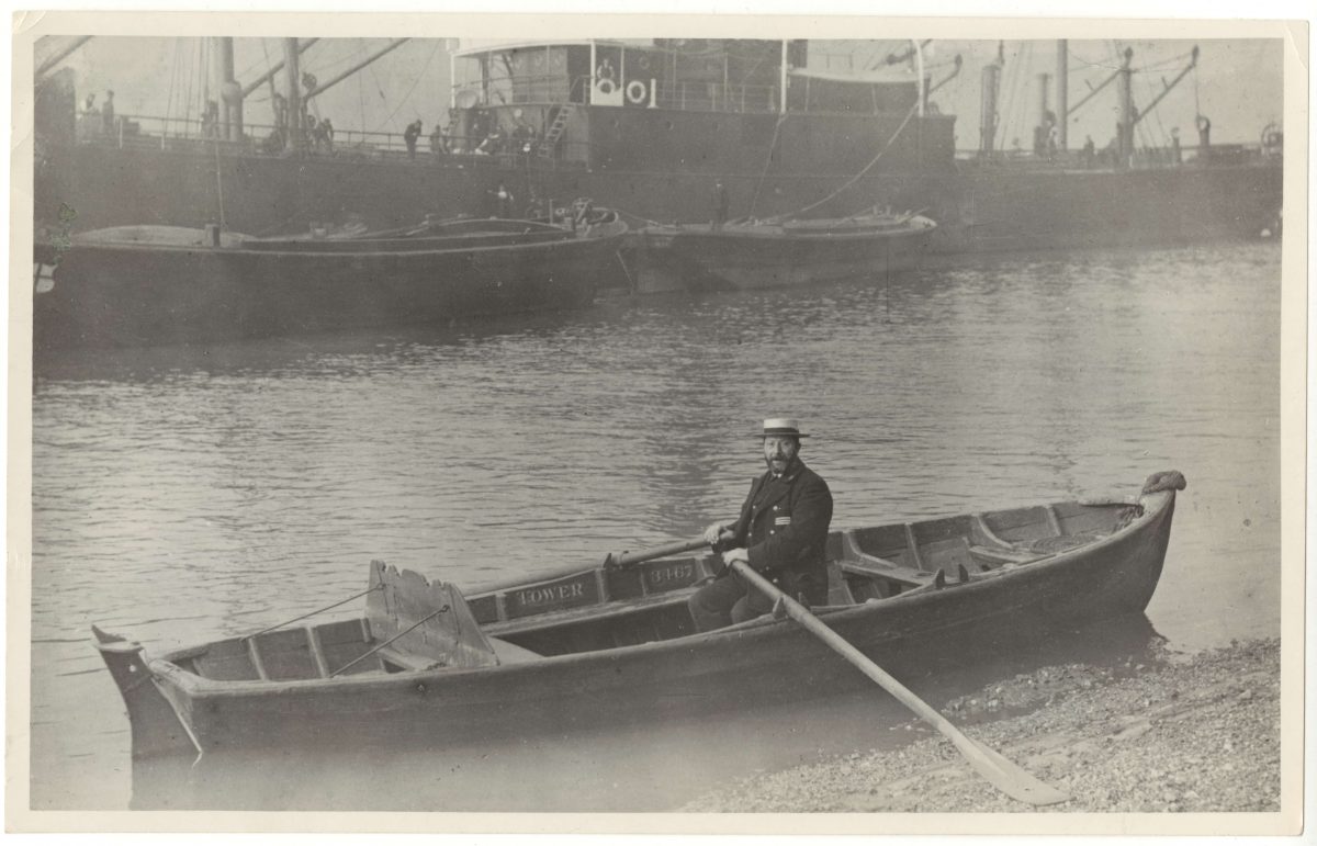 This is a very old, black and white photograph of a middle-aged bearded man sat in a long wooden boat, known as a skiff. He wears a dark uniform and a straw boaters hat. He has a long wooden oar in each hand and is looking directly at the camera. 