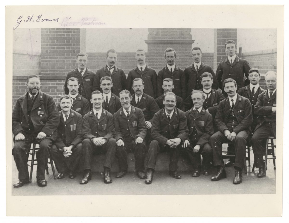 All 20 men in this black and white photo have facial hair, except two, the youngest looking men. The first two rows of men are sat down, the back row stands up. All look at the camera with varying degrees of happiness on their faces. George Evans looks to be the heaviest set man of the lot, the rest are fairly slight. They all wear postal uniforms; trousers, a tie and waistcoat, a thick jacket and shiny black shoes. 