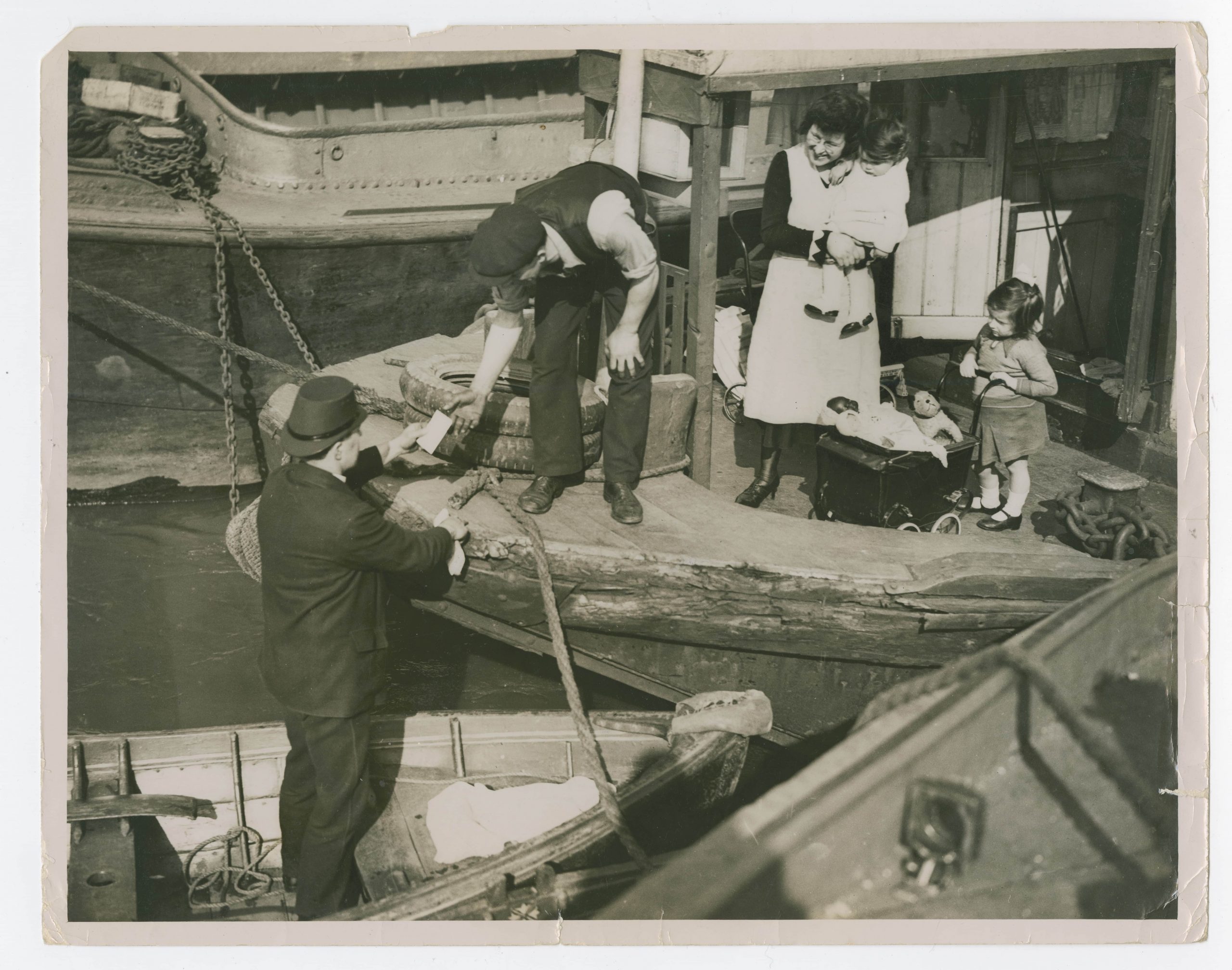 Herbert stands in the skiff, which is oared to a dock by a thick rope, handing a letter to a man, stood next to his young family. The woman stood next to the man, bending over to grab the letter, wears a white smock and is carrying a little girl, also in a dress. An older little girl stands on the far right of her family holding her toy pram.
