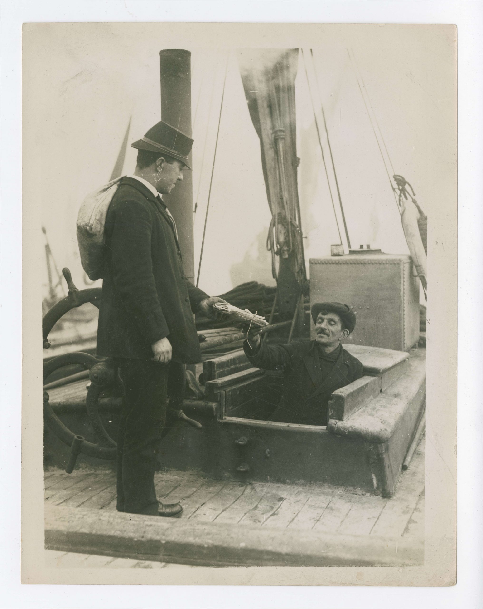 Herbert, wearing his uniform and white sack slung over his shoulder, hands a stack of letters to a man, popping up from below deck of his ship. You can see the steam from the ship's engine coming out of the chimney on the boat.