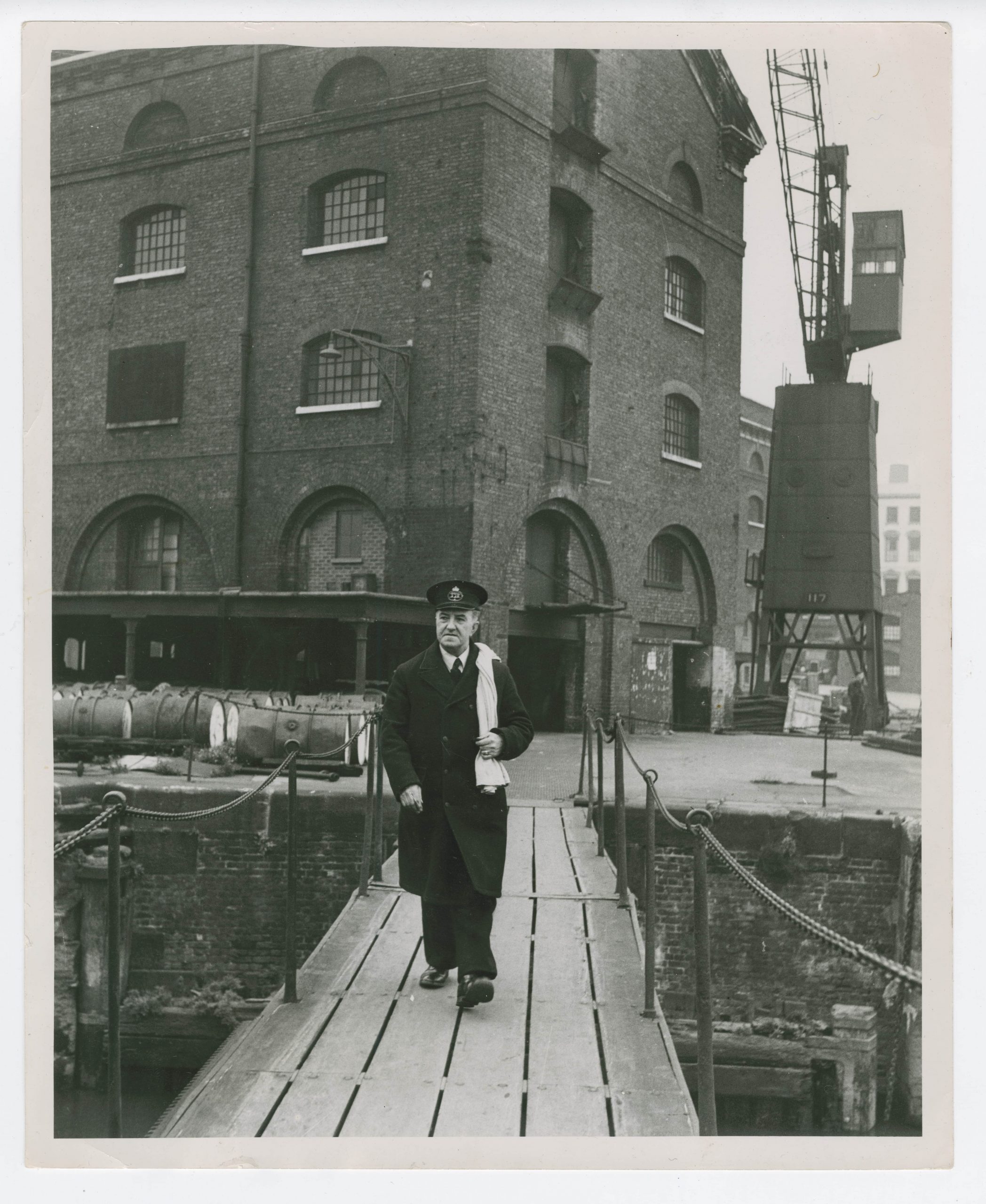 Herbert looks much older in this image than he does in the previous. An older, larger Herbert, with greying hair once again has his trusty white sack slung over his shoulder. He is walking along a wooden bridge, which leads away from a large industrial warehouse. The bridge goes over the Thames.