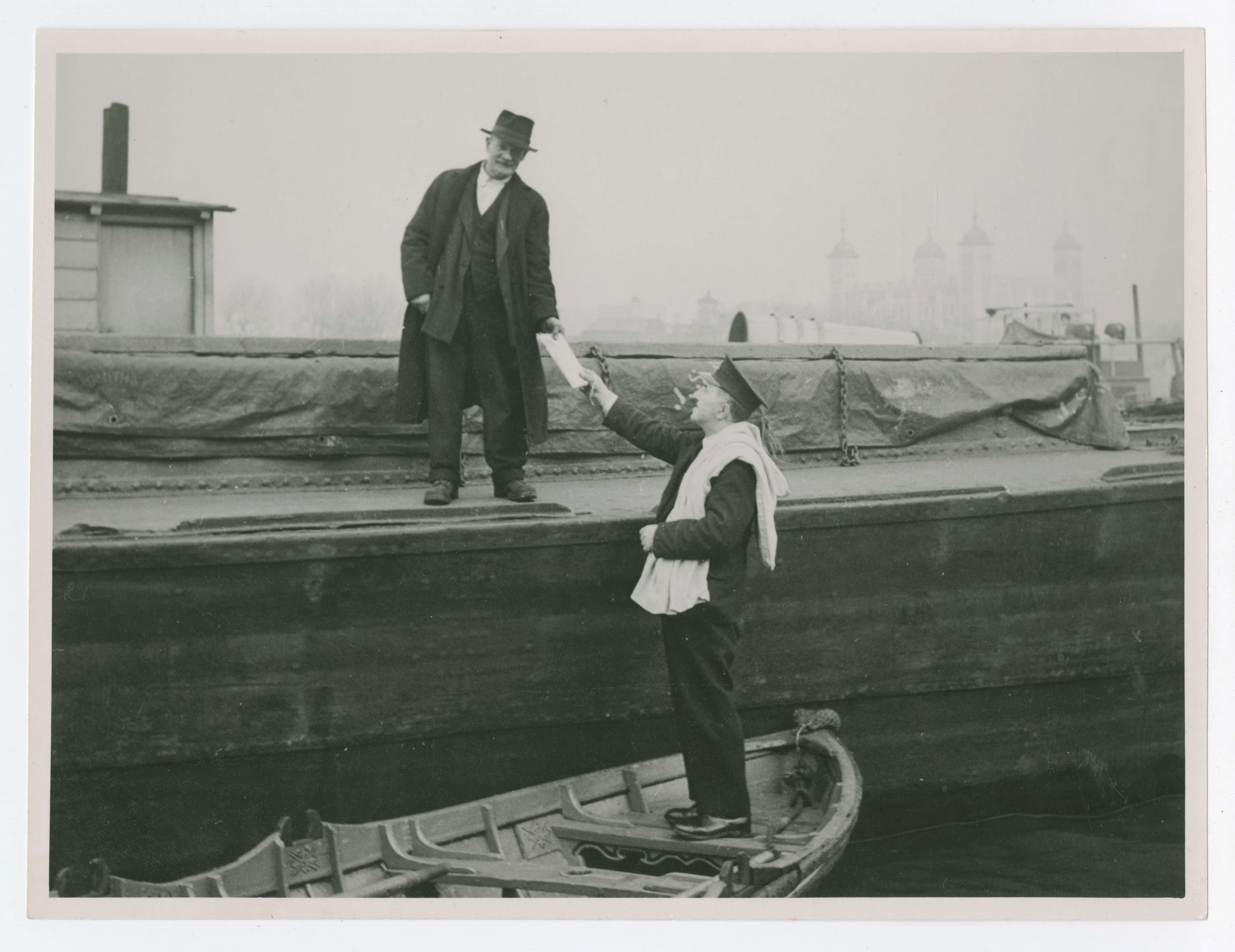 Herbert stands on one of the seats of his skiff, handing a white envelope up towards a man stood on the dock. Herbert is wearing his all black uniform, with postman cap, with a white sack tossed over his shoulder. The man he is handing the letter to looks directly at the camera and wears baggy looking dark clothes, a hat and a long trench coat.