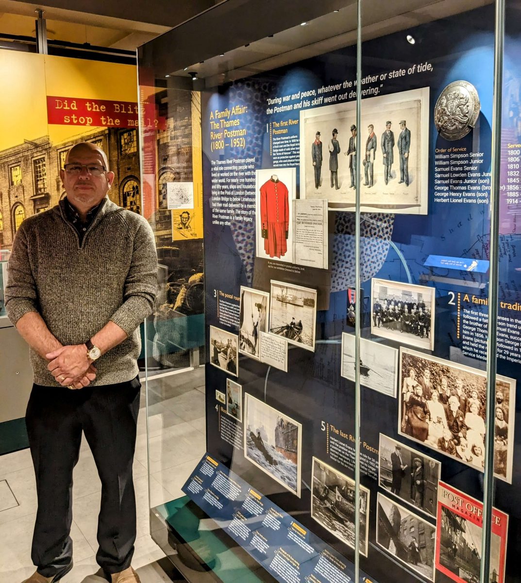 A middle aged, stocky and tall, white man wearing glasses stands in front of a museum display case. He is dressed in smart-casual attire with a zip up fleece, black suit trousers and comfy shoes on.