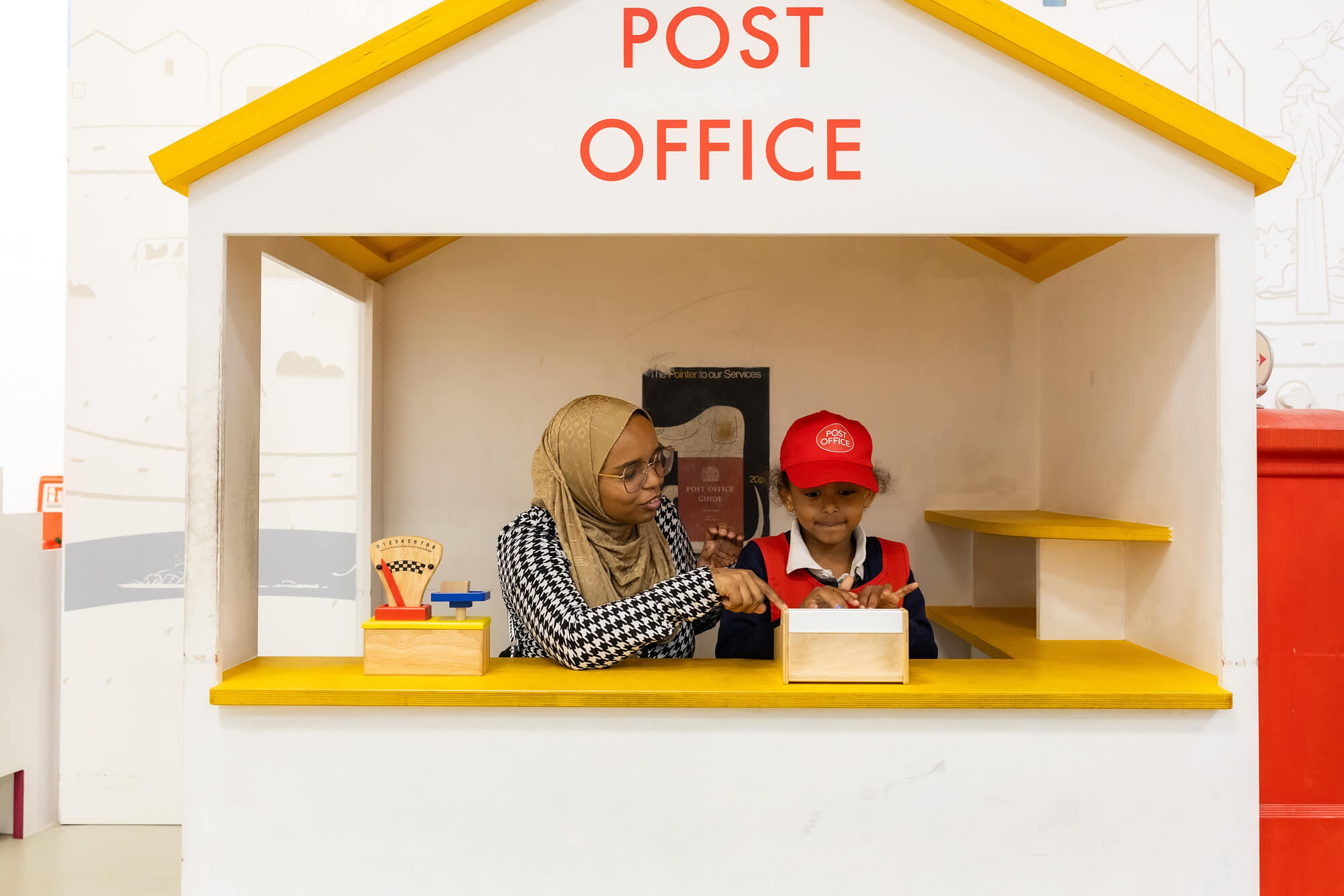 A teacher plays with her pupil in our miniature Post Office - a white shed with yellow trip. It says 'Post Office' in red font near the roof. The school child wears a red overall and red cap, as she is playing dress up. Her school uniform of a white polo shirt and blue jumper are peaking out.