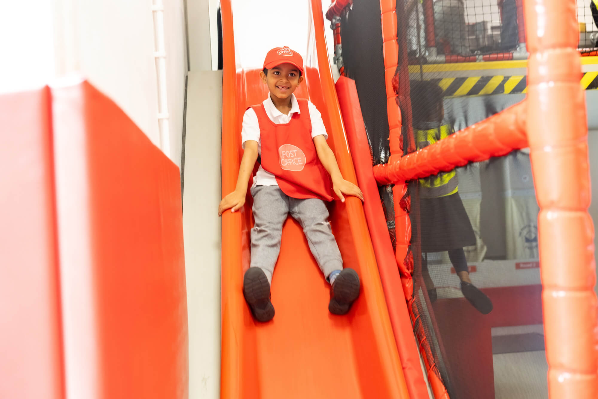 A young smiling boy slides down a bright red slide, legs first. He is wearing a red bib and red hat, grey trousers and a white polo shirt.