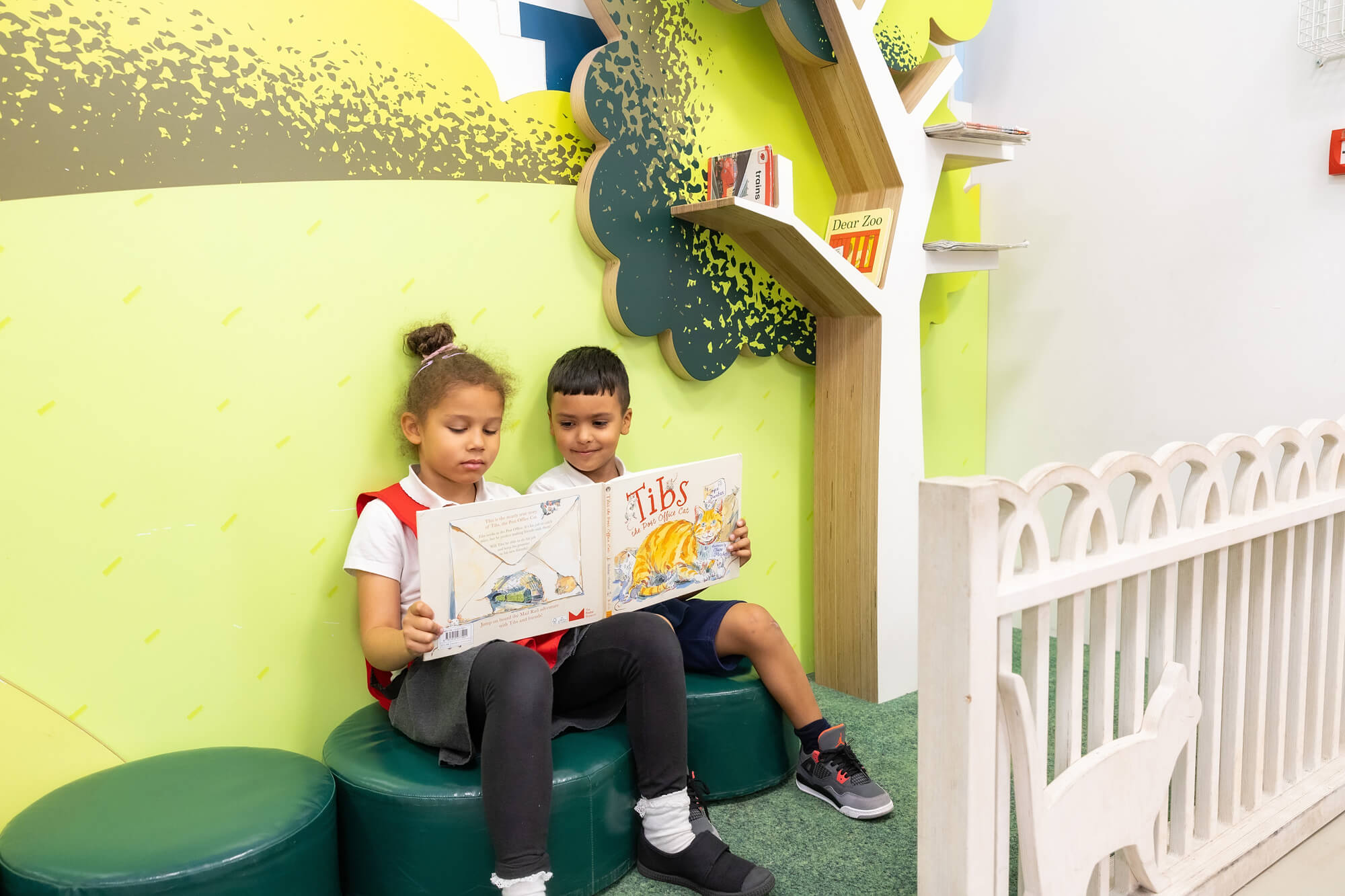 A young boy and girl sit reading in a green space, sat on comfy green stools. The both hold a side of the large book each. The boy is laughing, the girl is concentrating.