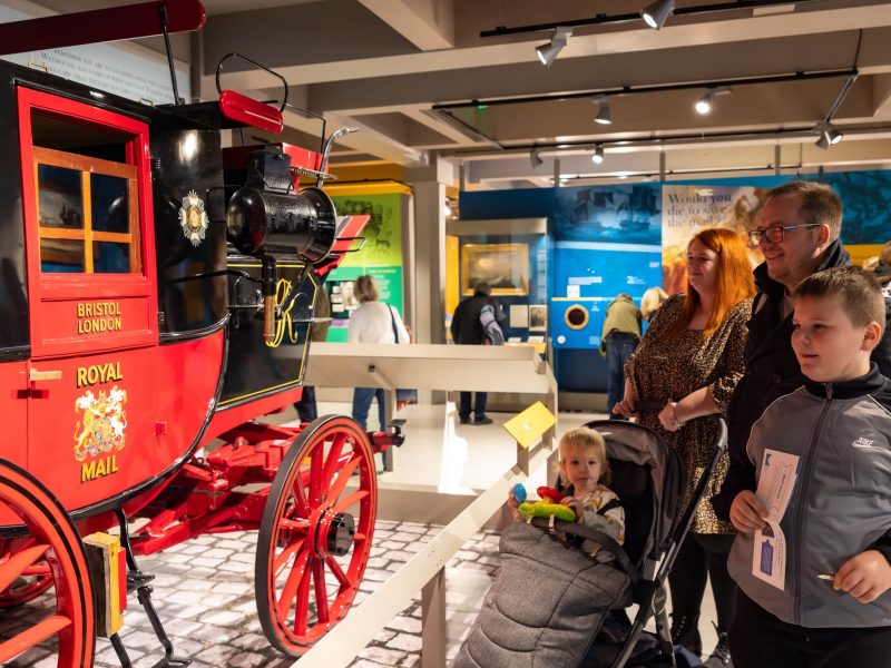 A mum and a dad look at the bright red mail coach with their young son and even younger daughter. The little girl is in a buggy.