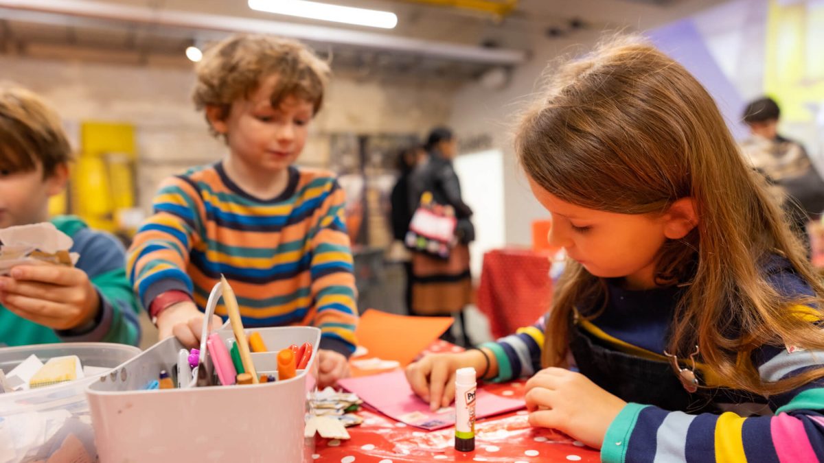 Three brown-haired children wearing long-sleeved t-shirts taking part in an arts and crafts activity. They are holding Pritt Sticks and pencils.