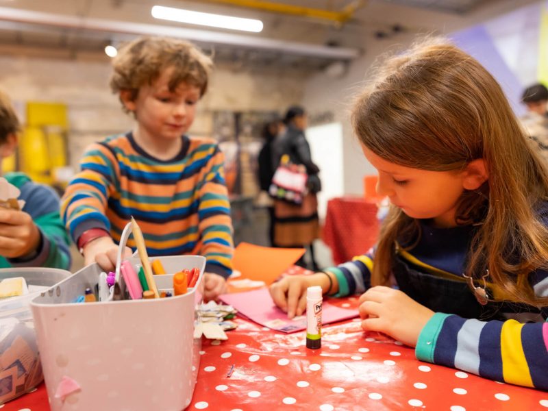 Three brown-haired children wearing long-sleeved t-shirts taking part in an arts and crafts activity. They are holding Pritt Sticks and pencils.