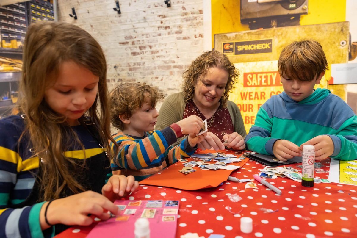 A mum, with blonde curly hair, sits at her table with two fair haired boys and a browned haired girl. All of them are sticking stamps down to a piece of card.