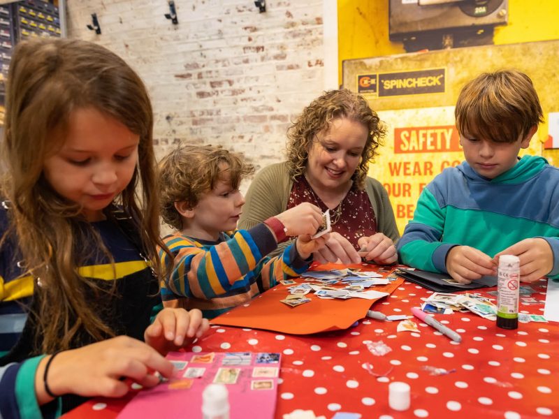 A mum, with blonde curly hair, sits at her table with two fair haired boys and a browned haired girl. All of them are sticking stamps down to a piece of card.