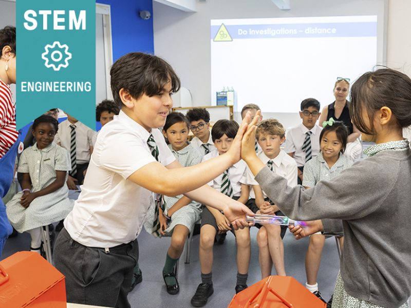 Skills: Stem, engineering. Two school children play a game while others look at them.