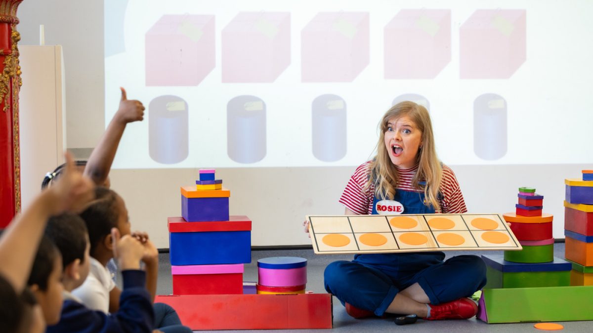 A learning facilitator is sitting on the floor surrounded by children. She holds a game and looks at them with a surprise face.