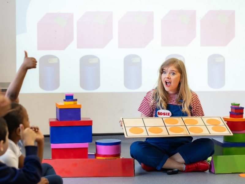A learning facilitator is sitting on the floor surrounded by children. She holds a game and looks at them with a surprise face.