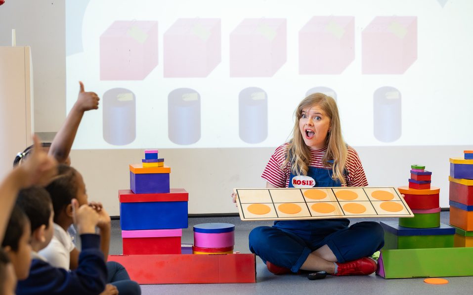 A learning facilitator is sitting on the floor surrounded by children. She holds a game and looks at them with a surprise face.