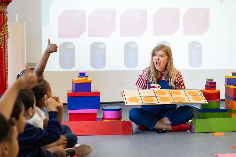 A learning facilitator is sitting on the floor surrounded by children. She holds a game and looks at them with a surprise face.