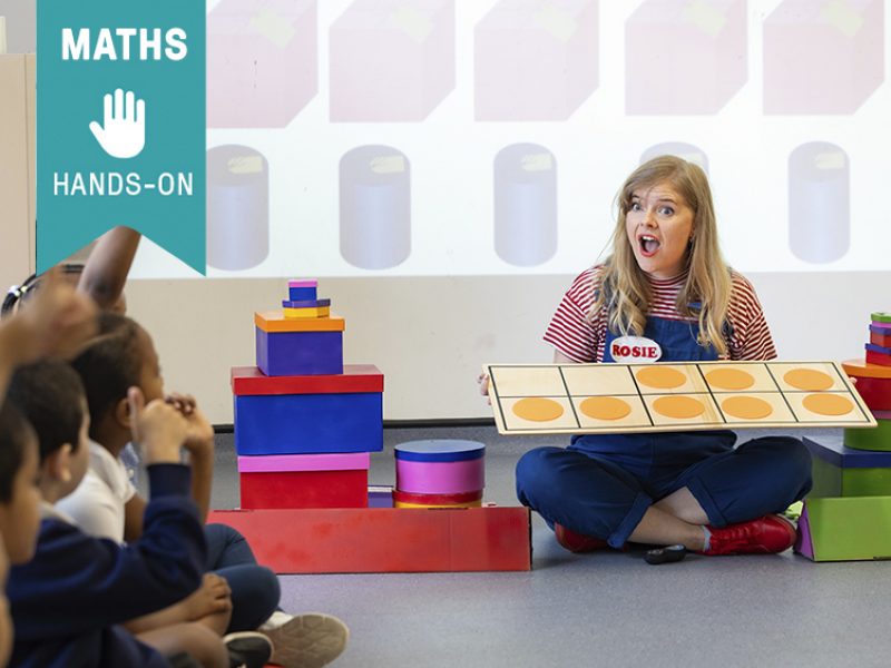 Skills: Maths, hands-on. Rosie holds a maths game while looking at school children sitting on a circle