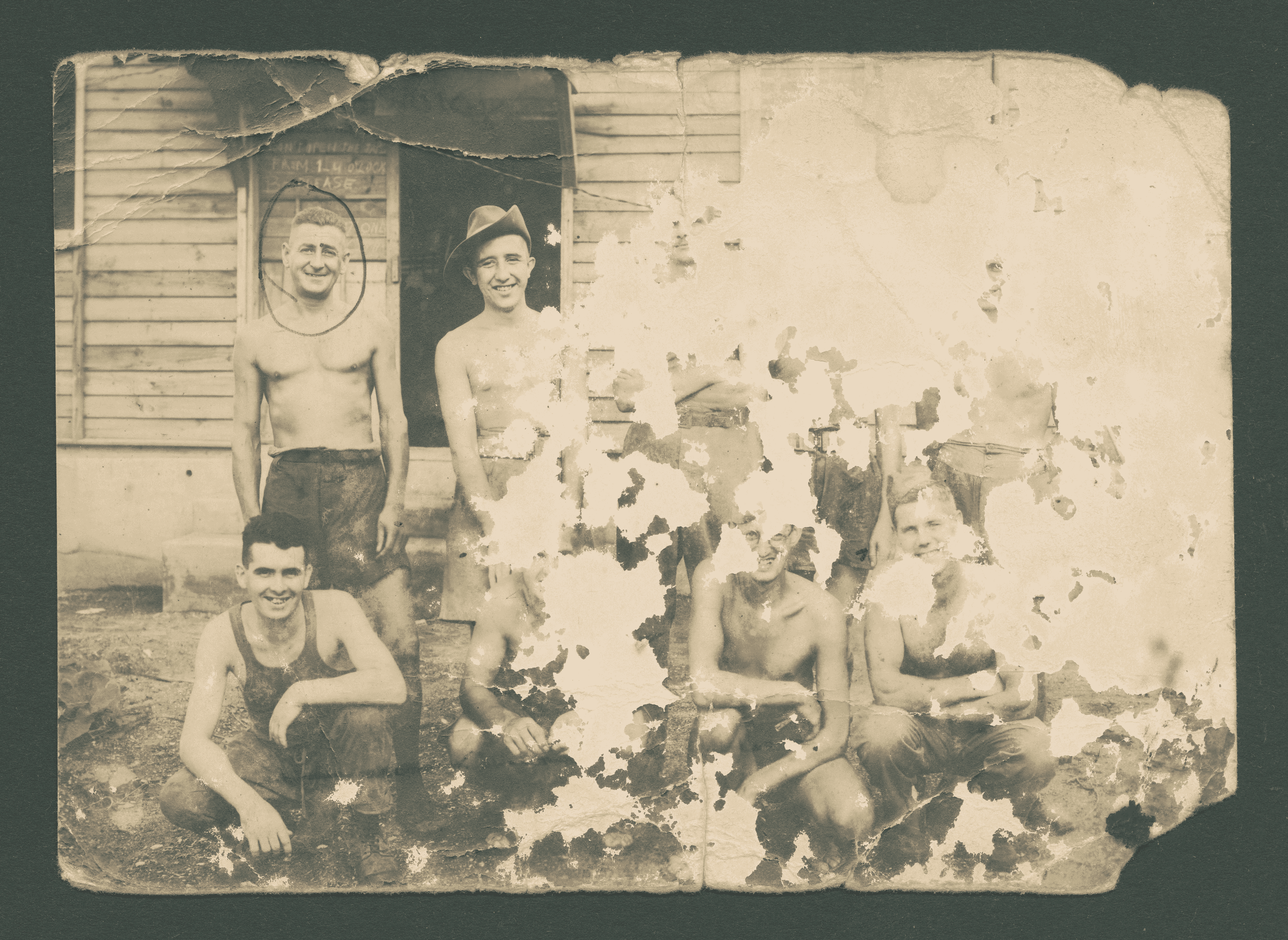 This sepia photo is so old and worn out that only 3 men are fully visible on it, out of 9. There is a row of men standing and a row below them crouching down. All men look directly to the camera smiling. They are either shirtless or wearing vests. They are all thin, their clothes don't fit them well.