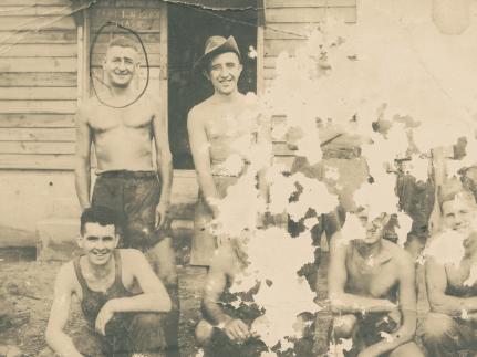 This sepia photo is so old and worn out that only 3 men are fully visible on it, out of 9. There is a row of men standing and a row below them crouching down. All men look directly to the camera smiling. They are either shirtless or wearing vests. They are all thin, their clothes don't fit them well.