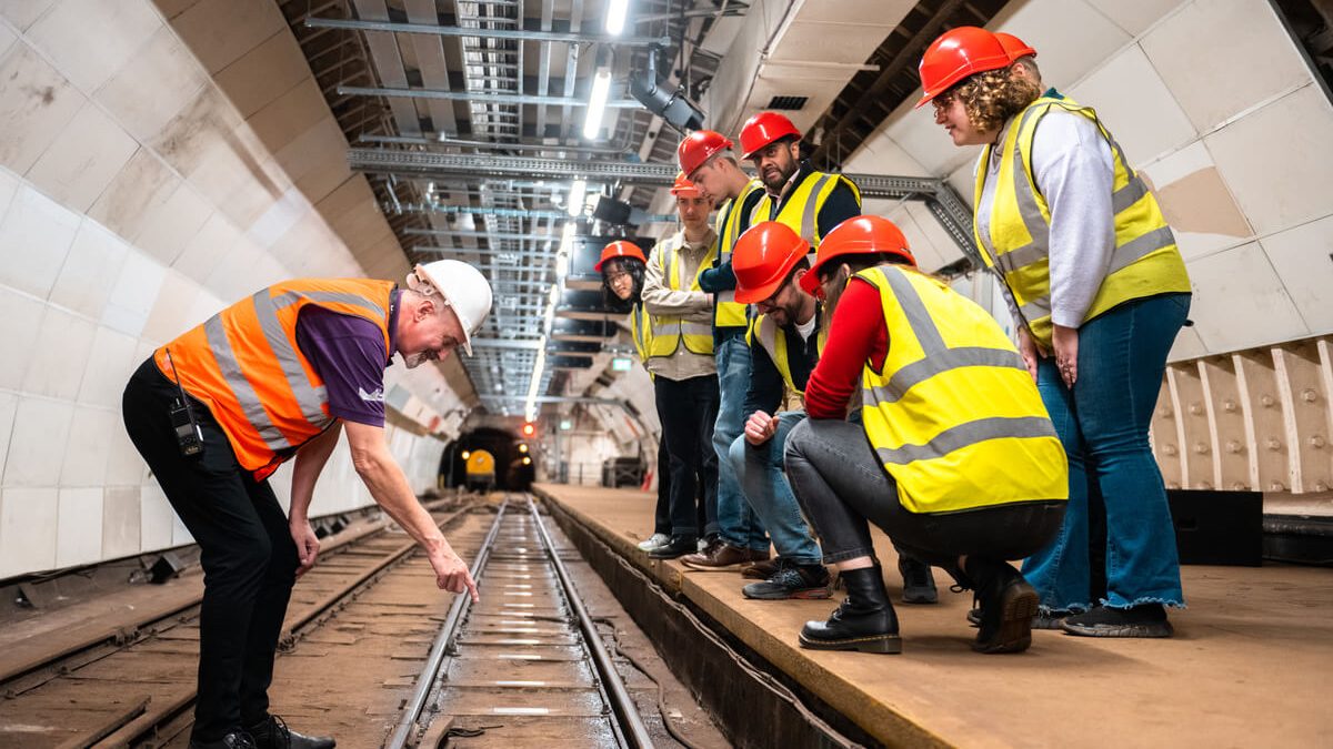 A group of people look down at a a train track from platform level. Next to the train track, a man points at a metal sleeper. All wear high vis vests and hard hats.