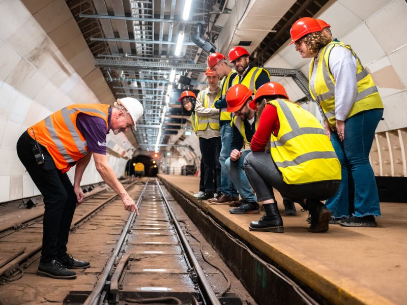 A group of people look down at a a train track from platform level. Next to the train track, a man points at a metal sleeper. All wear high vis vests and hard hats.