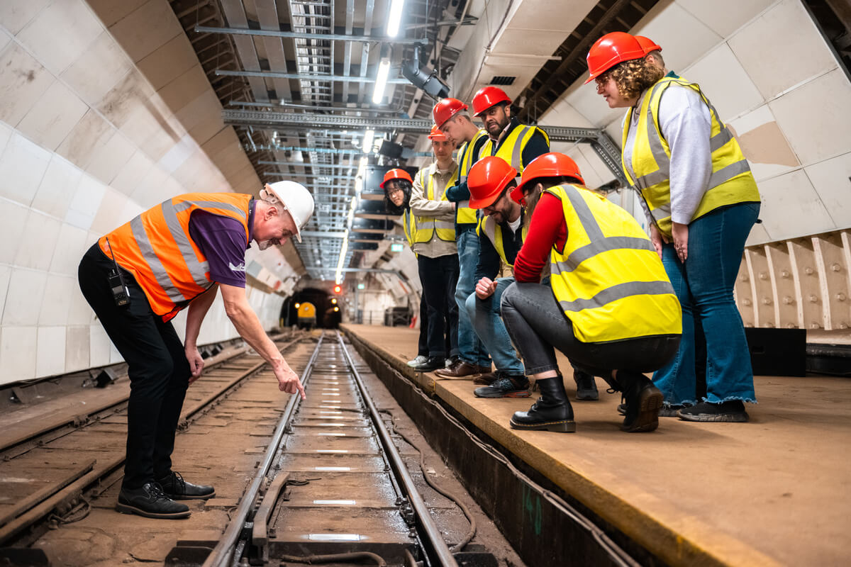 A group of people look down at a a train track from platform level. Next to the train track, a man points at a metal sleeper. All wear high vis vests and hard hats.