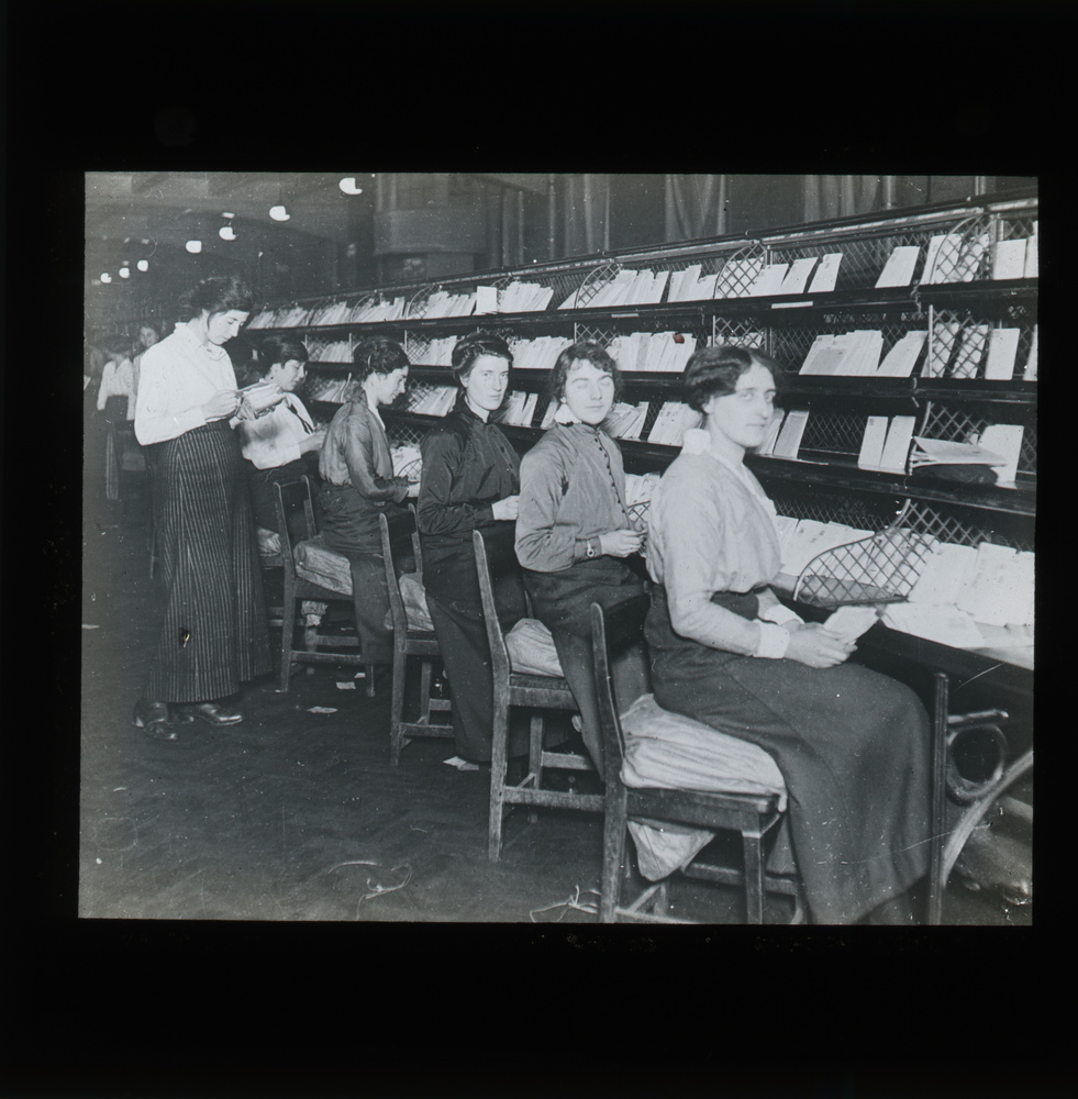 A group of women sitting in front of a long counter sorting mail. Some of them look at the camera.