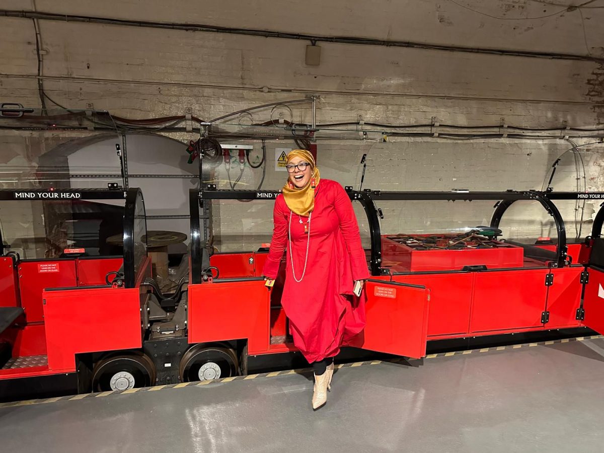 Onjali gets off the Mail Rail train while looking at the camera with a red smile. She's wearing a red dress and a yellow hijab.