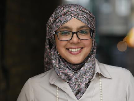 Onjali smiles to the camara. She's wearing glasses, a floral pattern hijab and a beige shirt.