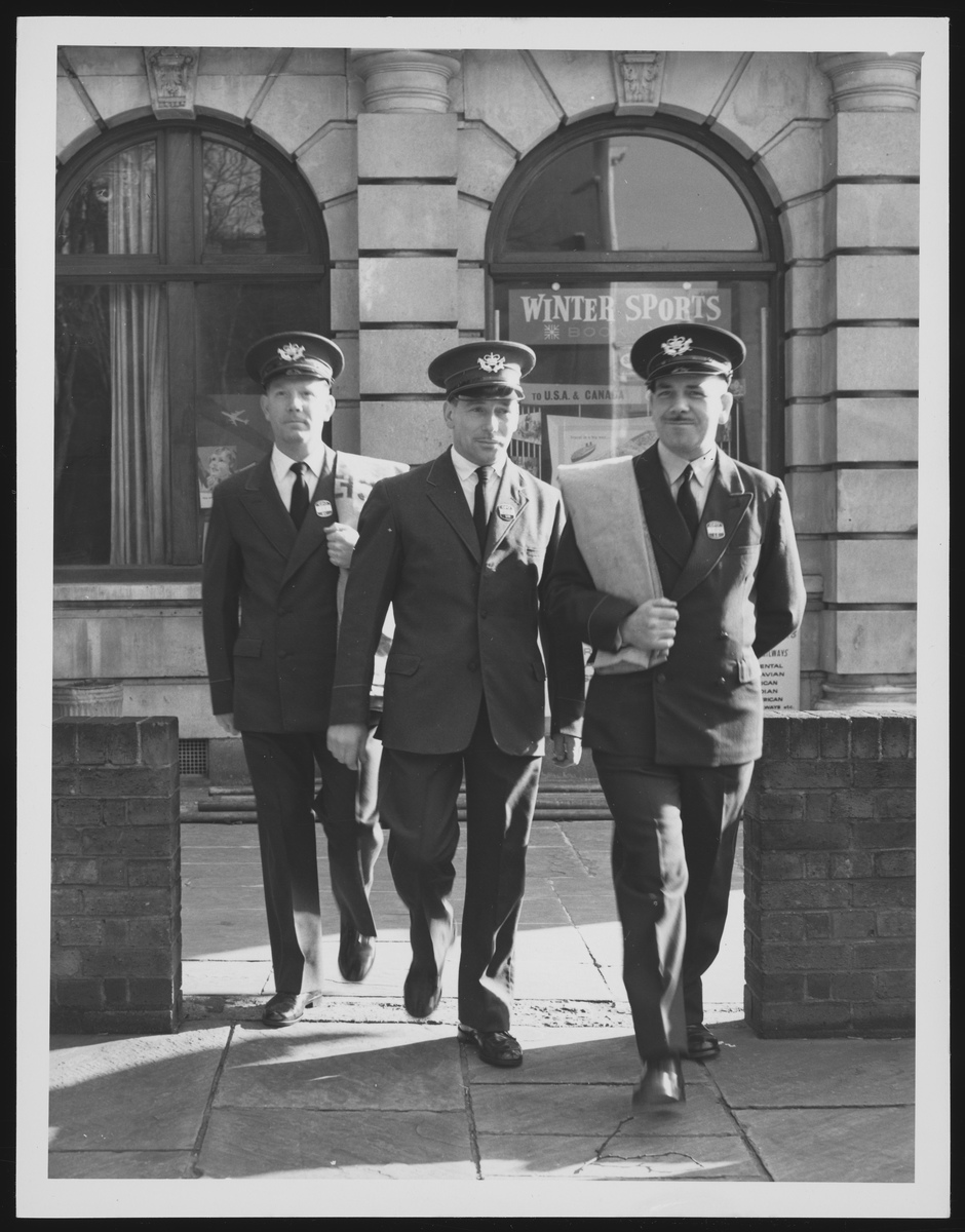 Black and white photograph of three white, male, postal workers. The men are wearing grey trousers and jacket with a white shirt and black tie. They are also wearing single peaked caps with a badge on each. The man at the front and the one at the back are carrying mail bags.
