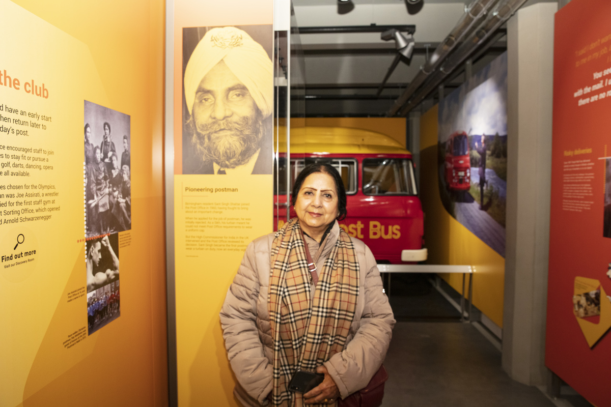 An Asian woman standing next to an interpretation panel with an image of Sant Singh Shattar on it. In the background there is a red and yellow post bus.