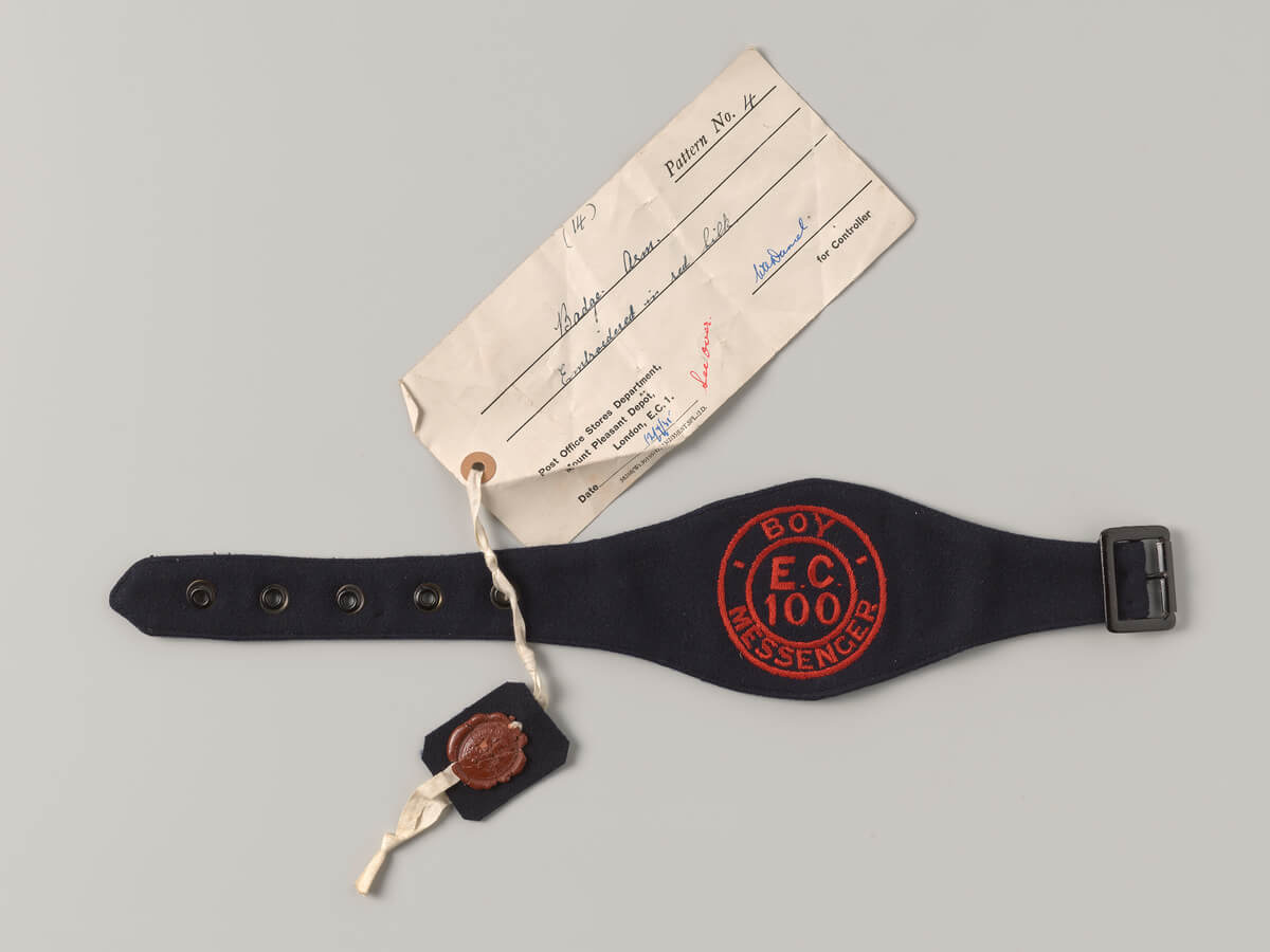 Blue armband with red embroidered text that reads ‘Boy Messenger E.C. 100’. This is within two concentric circles also in red. The armband has a metal buckle and five metal eyelets.