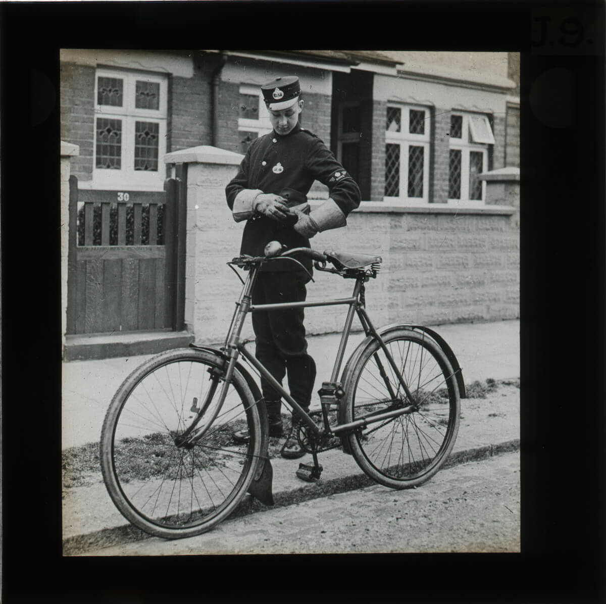 A Telegram Messenger next to his bike and in front of a house with the number 30 on its gate. He looks down at his pouch as he gets a telegram out of it.